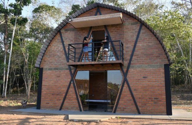 Edifício moderno de tijolos com telhado em arco, sacada e duas pessoas. Uma está com os braços erguidos. Rodeado por árvores.