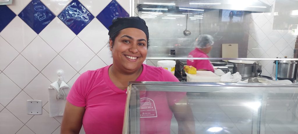 Mulher sorridente de camisa rosa e touca preta atrás de balcão de cozinha. Ao fundo, azulejos e funcionário.