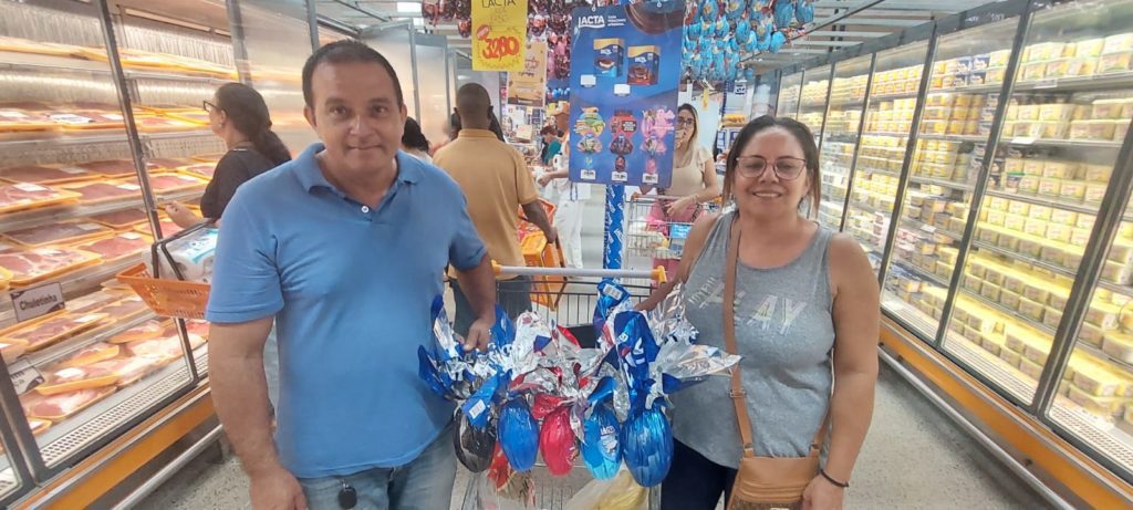 Duas pessoas sorrindo em supermercado com carrinho cheio de ovos de Páscoa coloridos.