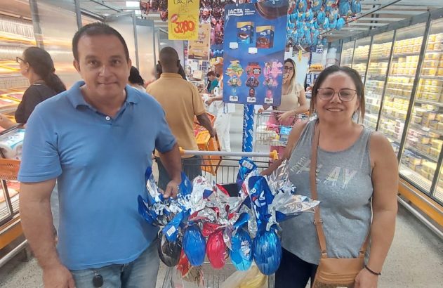 Duas pessoas sorrindo em supermercado com carrinho cheio de ovos de Páscoa coloridos.