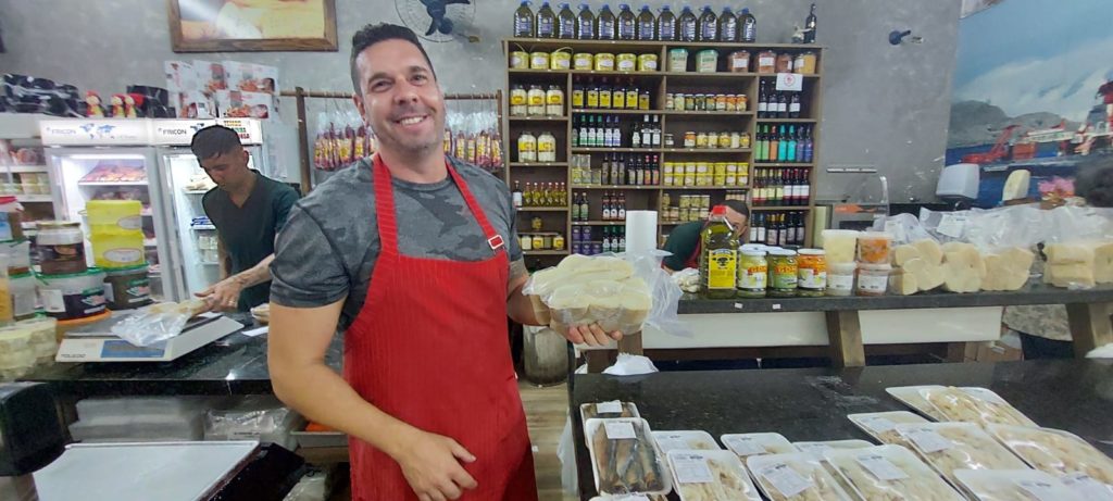 Loja de queijos: Homem de avental vermelho sorri atrás de balcão, outro funcionário trabalha no fundo.