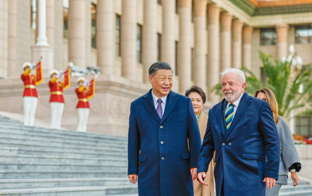 Presidentes Xi Jinping e Lula da Silva caminhando e sorrindo. Ao fundo, guardas de honra em frente a um edifício oficial.