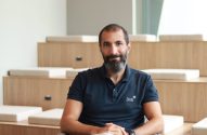 Homem barbudo com camisa polo Jive sorri, sentado em ambiente moderno com degraus de madeira.