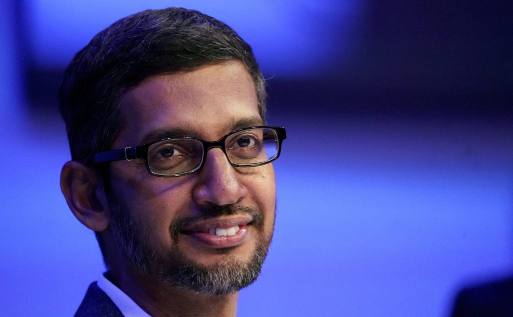 Sundar Pichai sorrindo, de óculos e barba, em fundo azul desfocado.