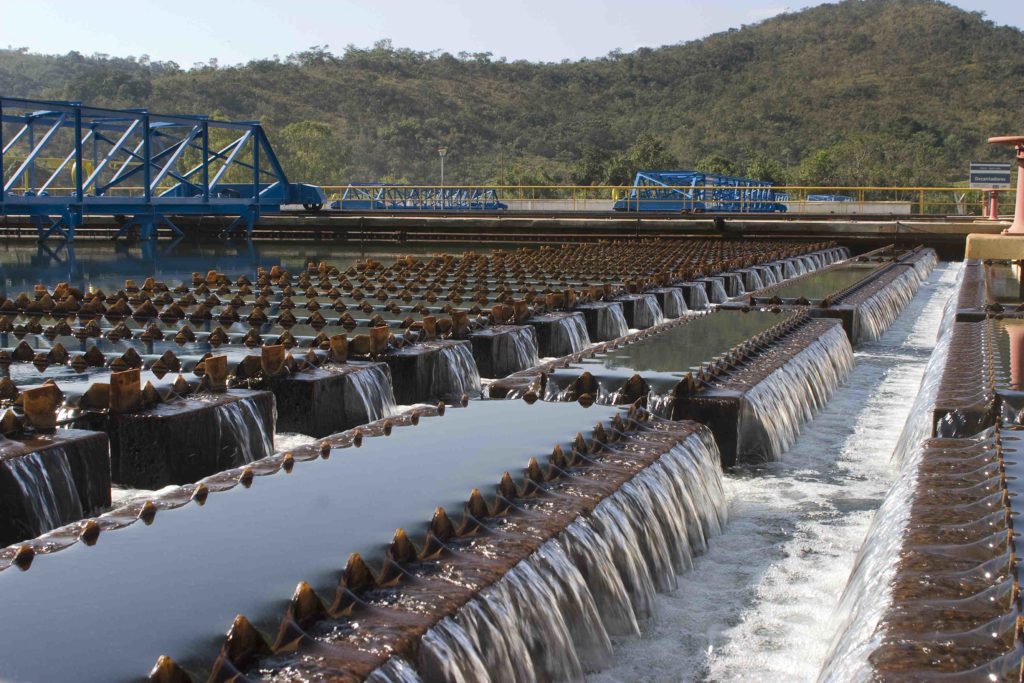 Estação de tratamento de água: fileiras de vertedouros com água cristalina caindo, estruturas azuis e paisagem montanhosa.