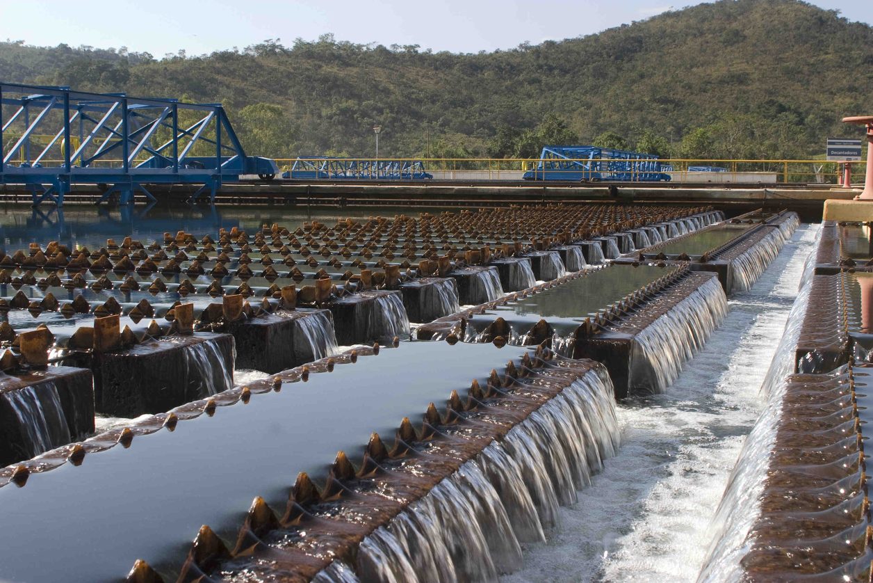 Estação de tratamento de água: fileiras de vertedouros com água cristalina caindo, estruturas azuis e paisagem montanhosa.