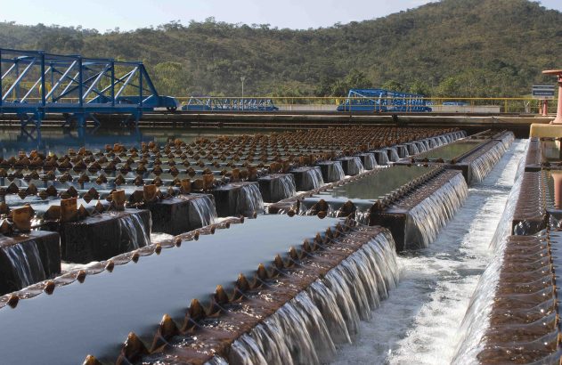 Estação de tratamento de água: fileiras de vertedouros com água cristalina caindo, estruturas azuis e paisagem montanhosa.