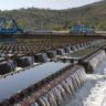 Estação de tratamento de água: fileiras de vertedouros com água cristalina caindo, estruturas azuis e paisagem montanhosa.