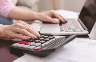 Mãos de uma mulher teclando em um notebook e fazendo contas em uma calculadora