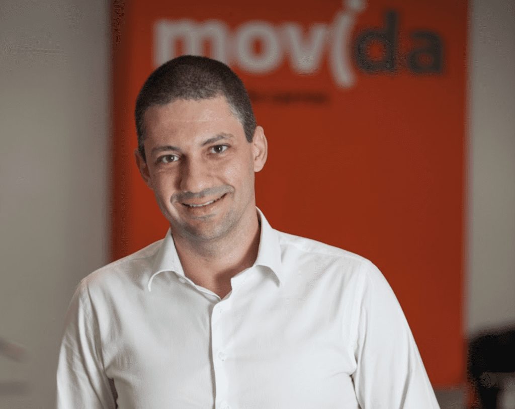 Homem sorrindo com camisa branca, logo da Movida ao fundo.