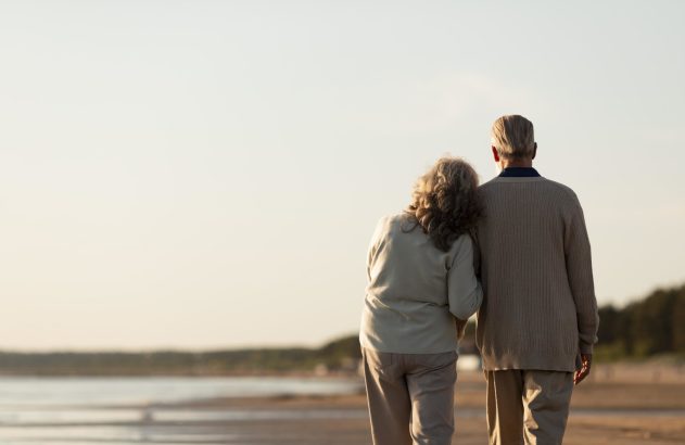 Casal idoso de costas andando na praia, mulher com a cabeça no ombro do homem.