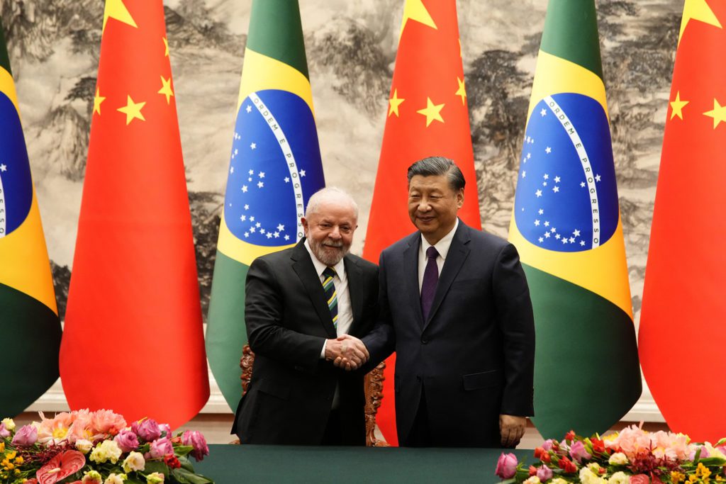 Presidente Lula e Xi Jinping apertam as mãos em frente a bandeiras do Brasil e da China.