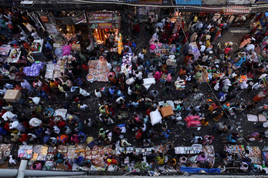Mercado lotado e movimentado, visto de cima. Multidão de pessoas e barracas com produtos diversos.