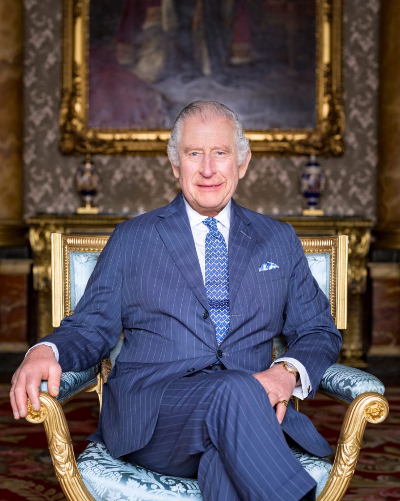 Rei Charles III sorri, sentado em poltrona ornada, com terno azul. Fundo clássico com quadro dourado.
