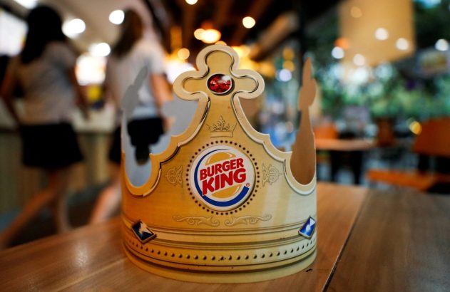 Coroa de papel do Burger King em mesa de madeira, com pessoas borradas e o interior de um restaurante ao fundo.
