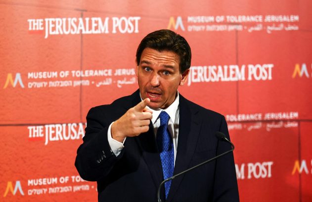 Homem de terno aponta o dedo e fala em frente a painel vermelho com logos 'Jerusalem Post' e 'Museum of Tolerance'.