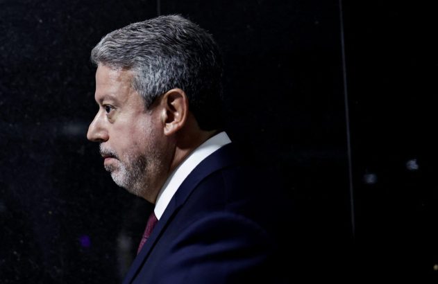 Homem com barba e terno em perfil, olhando para a esquerda em fundo escuro.