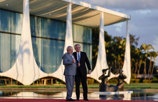 Lula e Alberto Fernández sorrindo e com polegares para cima, em frente a edifício governamental e espelho d'água.
