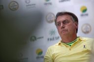 Jair Bolsonaro, camisa amarela com gola verde, com expressão séria. Fundo com logos.