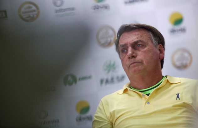 Jair Bolsonaro, camisa amarela com gola verde, com expressão séria. Fundo com logos.
