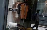 Vitrine de loja de roupas com placa "SALDOS", refletindo pedestre e manequim na rua.