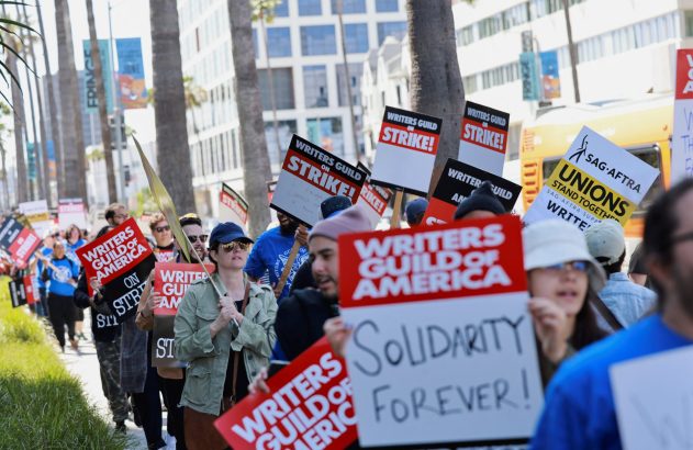 Manifestantes do Writers Guild em greve marcham com cartazes 'Writers Guild On Strike!' e 'Solidarity Forever!' na rua.