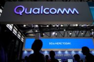 Silhuetas de pessoas em um estande da Qualcomm com seu logo roxo e letreiro '5G IS HERE TODAY'.