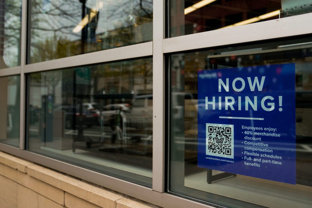 Cartaz 'AGORA CONTRATANDO!' em vitrine de loja, com QR code e informações sobre vagas de emprego.