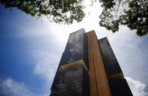 Prédio alto e escuro visto de baixo ângulo contra céu azul com sol forte e galhos de árvore.