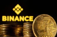 Moedas douradas de criptomoeda, incluindo uma com circuitos, em primeiro plano, com o logo da Binance ao fundo.