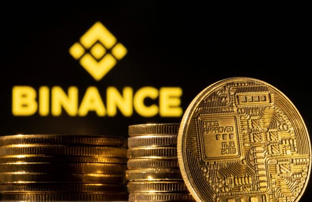 Moedas douradas de criptomoeda, incluindo uma com circuitos, em primeiro plano, com o logo da Binance ao fundo.