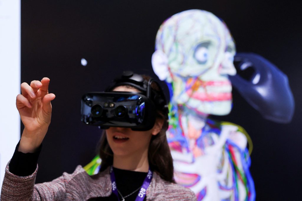 Jovem com óculos VR e mão levantada interage com um modelo 3D de esqueleto em tela.