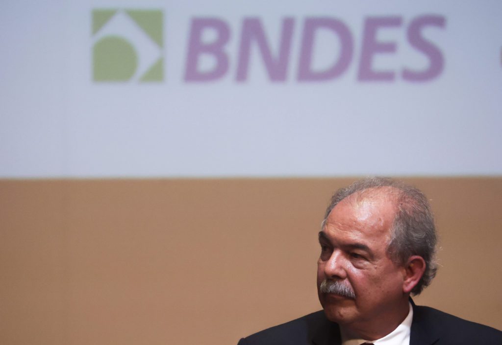 Homem de bigode em terno, olhando para o lado, com logo "BNDES" desfocado ao fundo.