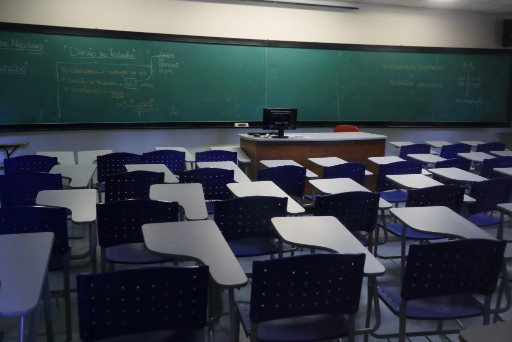 Sala de aula vazia com quadro verde, anotações de economia e carteiras azuis e brancas.