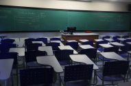Sala de aula vazia com quadro verde, anotações de economia e carteiras azuis e brancas.