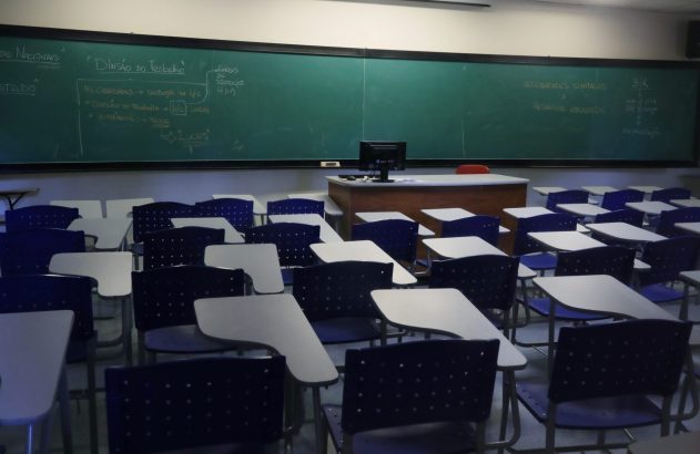 Sala de aula vazia com quadro verde, anotações de economia e carteiras azuis e brancas.