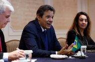 Homem de terno azul, Fernando Haddad, fala em mesa de reunião com outros participantes. Bandeira do Brasil na mesa.