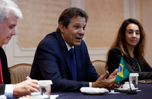 Homem de terno azul, Fernando Haddad, fala em mesa de reunião com outros participantes. Bandeira do Brasil na mesa.