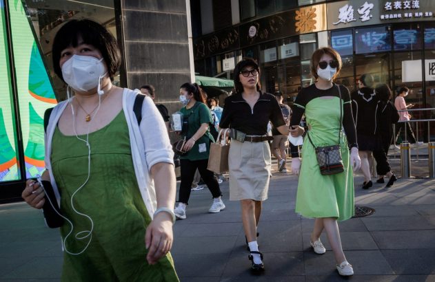 Pessoas com máscaras caminham em calçada movimentada de cidade, com prédios e lojas ao fundo.