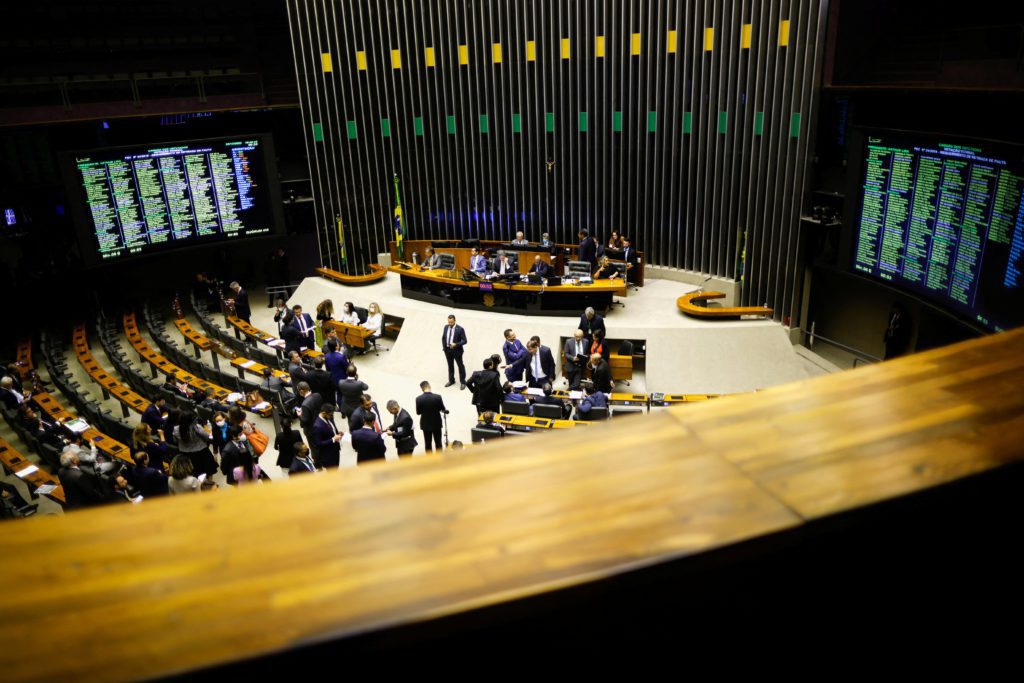 Vista aérea do plenário da Câmara Brasileira com parlamentares em discussão e movimento. Telas mostram votação.