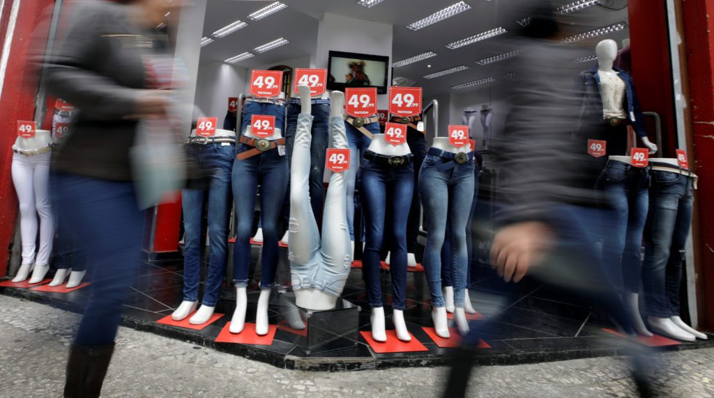 Manequins de jeans e blusas escuras em vitrine. Pessoas borradas passam em rápido movimento.