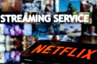 Smartphone com logo Netflix. No fundo, tela com 'Streaming Service' e vídeos borrados.