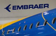 Logo azul da Embraer em parede branca, com parte da fuselagem de um avião azul e amarelo em primeiro plano.