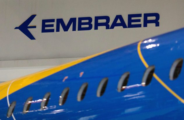 Logo azul da Embraer em parede branca, com parte da fuselagem de um avião azul e amarelo em primeiro plano.