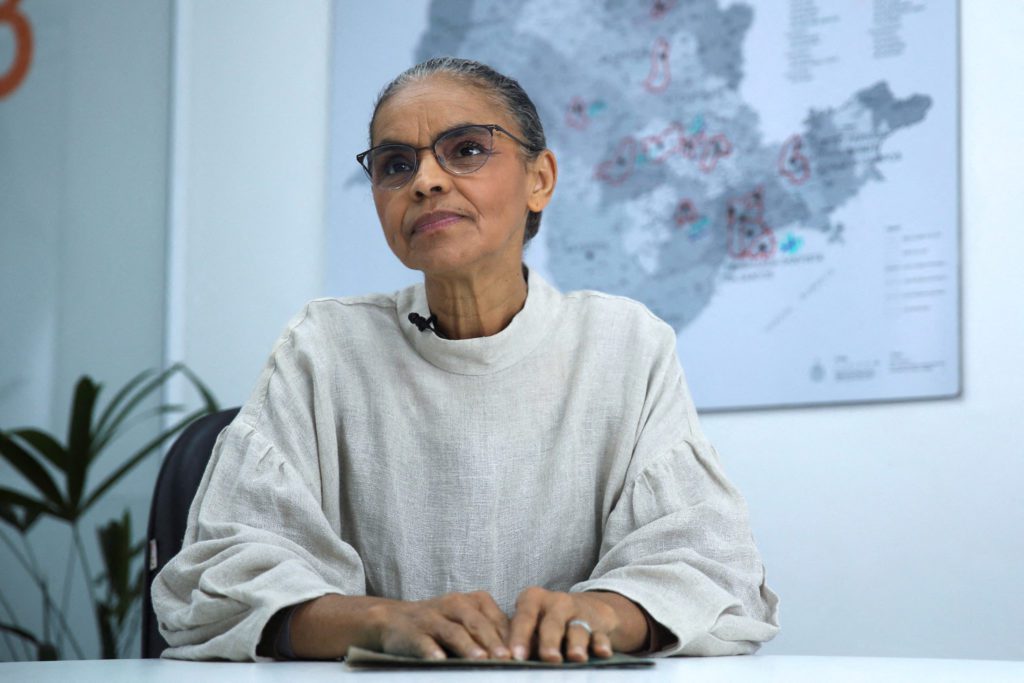Mulher de óculos sentada à mesa com microfone, mãos postas e um mapa ao fundo.