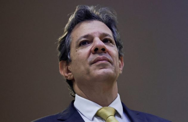 Homem de terno escuro, camisa branca e gravata amarela olha para o alto, com fundo desfocado.