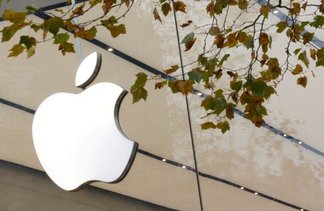 Logotipo iluminado da Apple em fachada de vidro, com galhos de árvore e folhas de outono.