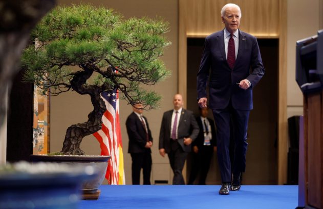 Presidente Joe Biden caminha para frente em evento. À esquerda, um grande bonsai e a bandeira dos EUA. Seguranças ao fundo.