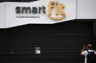 Fachada da academia Smart Fit fechada. Homem fotografa aviso na persiana.