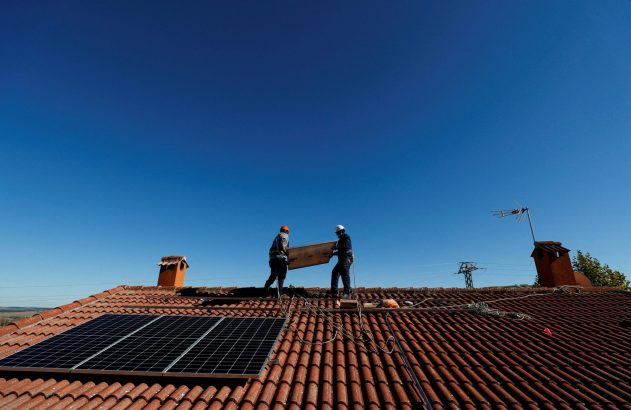 Dois trabalhadores instalam painéis solares em telhado de telhas, sob céu azul.